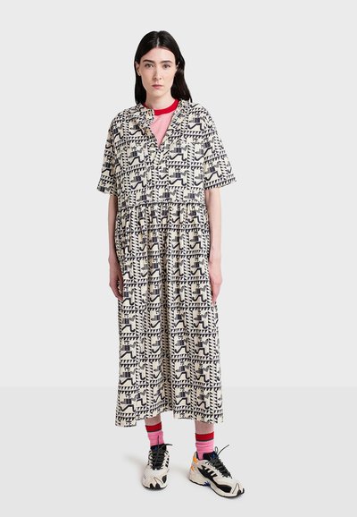 Midi-jurk met kraag, crèmekleurig met zwarte geometrische patronen, korte mouwen, gelaagd ontwerp, gedragen over een rood T-shirt en gecombineerd met zwarte sneakers.