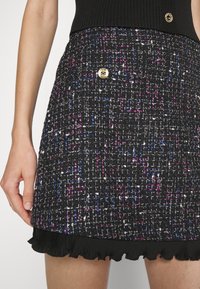 Jupe mini en tweed noir avec un design texturé agrémenté de fils colorés en violet et bleu, accentuée par un bouton doré et un ourlet froncé.