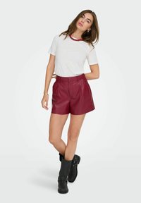 Burgunderfarbene Leder-Shorts mit hoher Taille und Falten, kombiniert mit einem weißen T-Shirt mit kurzen Ärmeln und schwarzen kniehohen Stiefeln. Rote Perlenkette.
