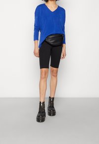 Femme portant un haut bleu à manches longues avec un col en V, un short de cycliste noir, un sac banane en cuir noir et des bottes de combat noires à semelle plateforme.