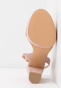 Sandale beige en daim à talon haut vue par le dessous, mettant en avant la semelle et le talon bloc sur un fond blanc uni.