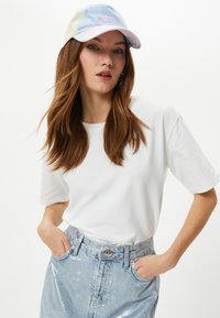 Vit t-shirt med kort ärm kombinerad med en ljusblå glansig denimkjol. En pastellfärgad tie-dye-keps har broderat "Men Sal Love" på framsidan.