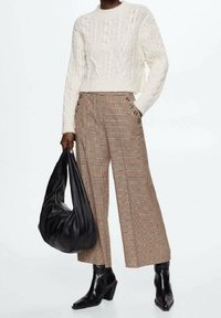 Geknipte crèmekleurige kabelgebreide trui, hoogliggende geblokte broek met knoopaccenten, zwarte tas en zwart leren laarzen met puntige neus.