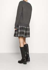 Pull en maille gris, jupe plissée à carreaux noir et blanc, et bottes noires montantes. La tenue présente une coupe décontractée et des matériaux texturés.