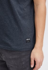 Mørkegrå bomulds T-shirt med korte ærmer, der har et tekstureret materiale og et lille sort mærke i kanten med hvid tekst.