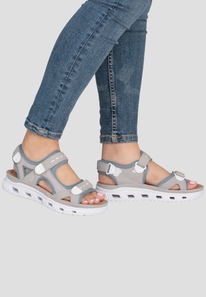 Pieds portant des sandales réglables grises et blanches associées à un jean denim bleu sur un fond neutre.