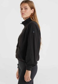 Veste en polaire noire avec col haut, poignets élastiques et fermeture éclair. Présente un petit logo sur la manche et une coupe décontractée.
