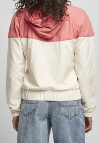 Lekki kurtka zewnętrzna o różowej górnej części i kremowej dolnej części. Posiada elastyczny dół i kaptur. Noszona z jasnoniebieskimi dżinsami.