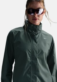 Moteris vilkinti tamsiai žalią Nike striukę su aukštu kaklu ir didelėmis atspindinčiomis sportinėmis akinių nuo saulės prieš vientisą foną.