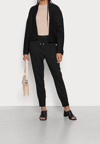 Czarny krótki kardigan, beżowy top, czarne spodnie dresowe i czarne buty na słupku z odkrytymi palcami. Modelka nosi jasnobeżową torbę.