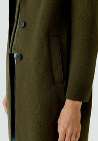 Manteau en mélange de laine verte avec une finition texturée, doté d'une fermeture à un bouton et d'une poche latérale, présenté avec un gros plan sur le détail du tissu.