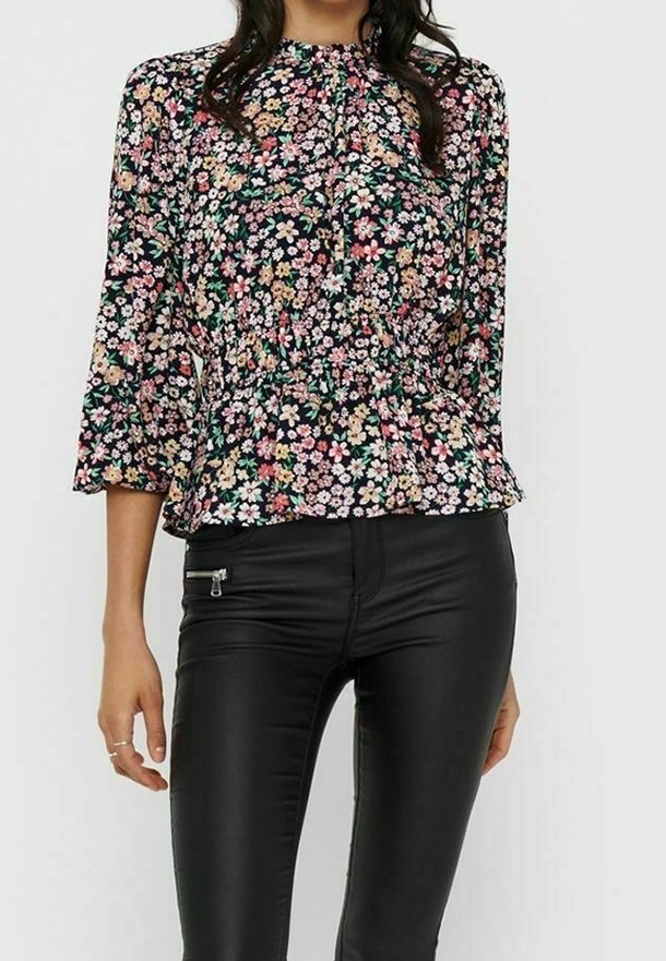 Femme portant un chemisier à manches longues à motif floral avec des fleurs roses, blanches et jaunes et un pantalon en cuir noir ajusté avec une poche zippée.