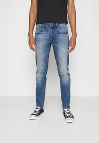 Jeans de corte slim en denim azul claro con un ligero desgaste, diseño de cinco bolsillos y detalles de costura. Combinados con zapatillas negras.