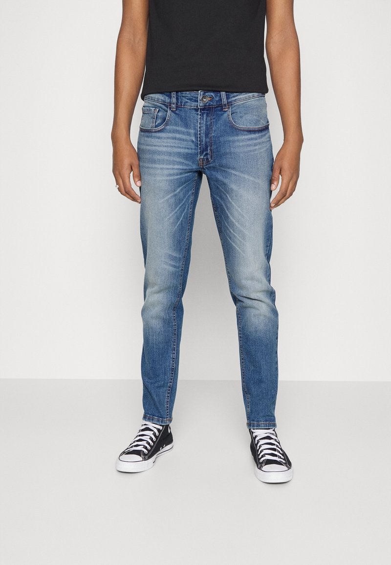 Jeans de corte slim en denim azul claro con un ligero desgaste, diseño de cinco bolsillos y detalles de costura. Combinados con zapatillas negras.