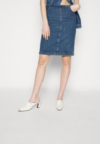 Džínová tužková sukně v tmavě modré barvě s hladkým povrchem a lehkým opotřebením, spárovaná s bílými sandály na vysokém podpatku s otevřenou špičkou a geometrickým vzorem.