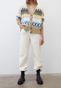 Cardigan en tricot avec des motifs géométriques en beige, bleu, jaune et marron ; présente un col en V, des boutons et des poignets côtelés sur un pantalon crème et des bottes noires.