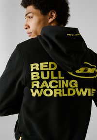 Sudadera negra con texto amarillo "RED BULL RACING WORLDWIDE" en la parte trasera. Cuenta con capucha ajustable con cordones y un bolsillo canguro. Fabricada con un tejido suave.