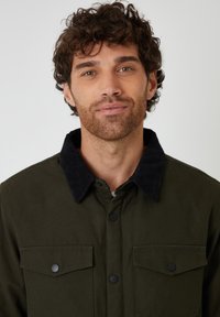 Ciemnozielona kurtka z czarnym kołnierzem z kordurowej tkaniny, z zapięciem na guziki z przodu i dwiema kieszeniami na piersi, wykonana z sztywnej tkaniny.