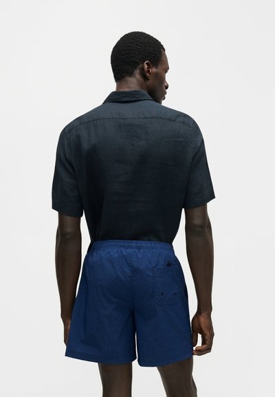 Άνδρας που φοράει σκούρο μπλε κοντομάνικη μπλούζα και μπλε ελαστικό σορτς με ελαστική μέση, γυρισμένος από πίσω προς ένα μονόχρωμο φόντο.