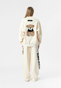 Sudadera oversized color crema con una aplicación de oso de peluche en la parte trasera, combinada con pantalones de pierna ancha a juego. Detalles en negro en el oso.