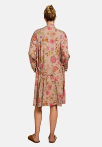 Beige stoffen gewaad met een bloemenpatroon, voorzien van veelkleurige geborduurde bloemen, lange mouwen en een strikceintuur in de taille.