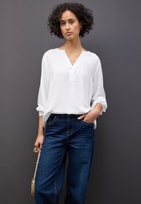 Blusa bianca a maniche lunghe con scollatura a V e pattina, abbinata a jeans dritti blu scuro. Sfondo grigio.
