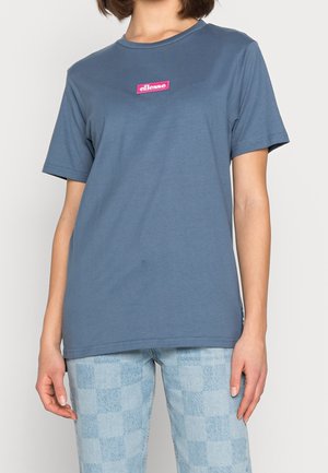 Persona con una camiseta azul con el logo "ellesse" en rosa en el pecho y jeans a cuadros azul claro, de pie contra un fondo liso.