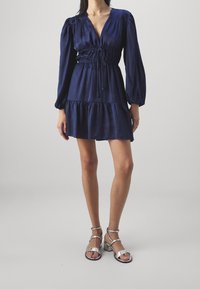 Námořnické modré šaty s dlouhými rukávy a hlubokým výstřihem do V, stažené v pase šňůrkou. Vícevrstvá sukně a hladký materiál. Stříbrné sandály na podpatku.