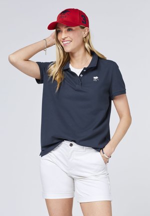 Marineblaues Poloshirt mit kleinem weißen Logo, kombiniert mit weißen, krempelbaren Shorts. Eine rote Baseballmütze mit gestickten Details und der Nummer 3.