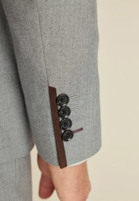 Manga de blazer de espiga gris con acento de tela marrón, que presenta cuatro botones negros y un detallado de costura en colores contrastantes.