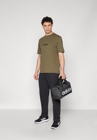 Zelené bavlněné tričko s černým logem Adidas, černé tepláky a černé tenisky s bílými akcenty. Drží černou tašku Adidas.