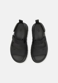 Svarte sandaler laget av gummi og stoff, med to justerbare stropper, en teksturert fotseng og en fleksibel såle.