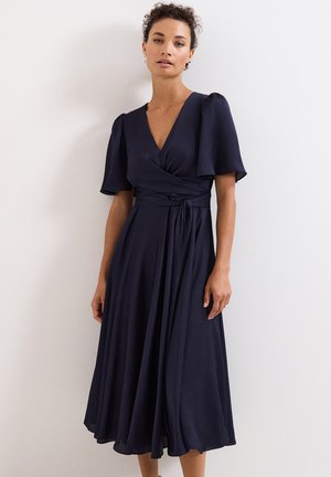 Vrouw die een marineblauwe wikkeljurk draagt met korte fladdermouwen en een gestrikte taille, staand tegen een effen witte muur.