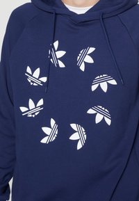 Marineblå hættetrøje med hvide blomsterlogo prints. Har en hætte med snøre og raglanærmer, lavet af blødt, tekstureret stof.