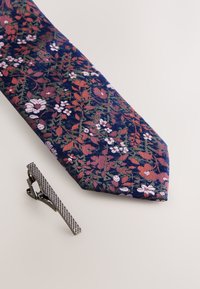Tmavě modrá květinová kravata s červenými a růžovými květy a zelenými listy; doplněná o stříbrnou sponu na kravatu s texturovaným povrchem a lineárním vzorem.