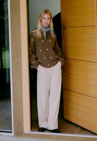 Bruine trui met witte stippen, beige wijde broek en een grijze sjaal. Draagt zwarte schoenen en staat naast een houten deur met glas.