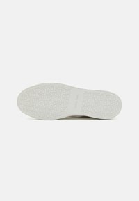 Hvit sneakersåle med et teksturert mønster som gjentar logoer og merkenavn, laget av gummi for grep og holdbarhet.