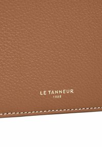 Materiale in pelle marrone con texture, con il nome del marchio "LE TANNEUR" e l'anno "1898" impresso in oro, caratterizzato da cuciture bianche lungo il bordo.