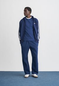 Tmavomodrá tepláková súprava s bielymi pruhmi, pozostávajúca z bundy na zips a zodpovedajúcich nohavíc. Nosí sa s priliehavým tmavomodrým tričkom a bielymi teniskami.