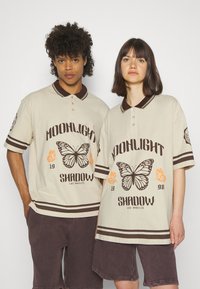 Beige polo shirts med brun krage og striper, med en sommerfugl-illustrasjon og teksten "MOONLIGHT SHADOW" i mørke og oransje detaljer.