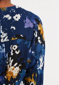 Vue de dos d'une personne portant un vêtement bleu foncé avec un motif floral en blanc, noir, violet et jaune moutarde.