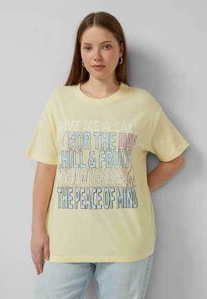 Junge Frau mit hellbraunen Haaren, die ein hellgelbes T-Shirt mit mehrfarbigem motivierendem Text trägt und vor einem grauen Hintergrund steht.