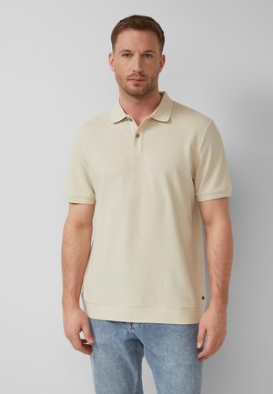 Man staat tegen een grijze achtergrond, draagt een beige poloshirt met korte mouwen en textuur, en lichtblauwe jeans, kijkt recht vooruit.