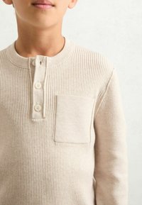 Beige geribde lange mouw top met een henley halslijn, een placket met drie knopen en een klein borstzakje. Zachte textuur, casual ontwerp.
