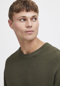 Pulover tricotat rib din lână verde măsliniu, cu guler rotund, având un material texturat și o croială relaxată, completat cu un detaliu de colier argintiu.