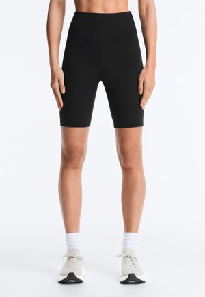 Femme portant un short de cycliste taille haute noir, des chaussettes blanches et des chaussures de course gris clair, debout les bras détendus le long du corps.