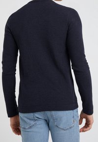 Man som bär en marinblå långärmad tröja och ljusblå jeans, sedd bakifrån mot en enfärgad ljus bakgrund.