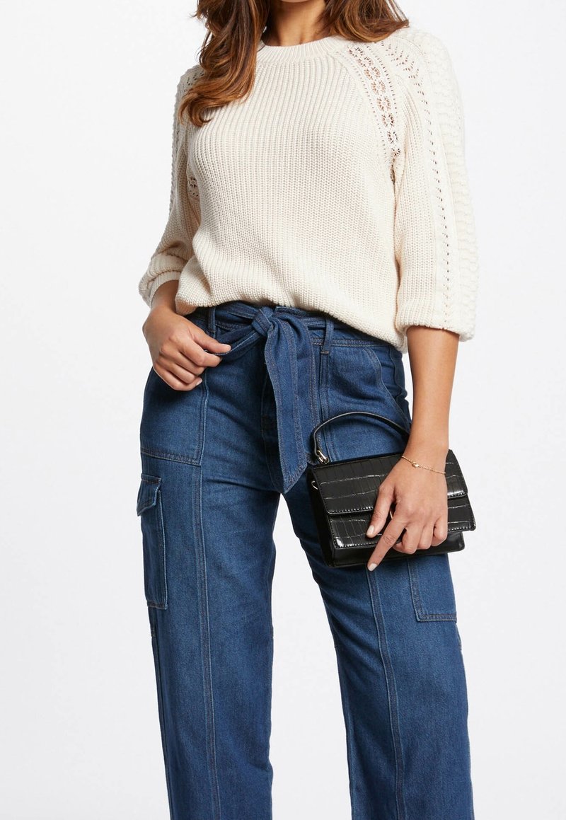 Frau trägt einen cremefarbenen Strickpullover mit Spitzenakzenten, hochgeschnittene blaue Denim-Hosen mit einem Taillengürtel, hält eine kleine schwarze Handtasche.