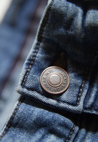 Denim z miedziowym guzikiem z napisem "JACK'S SPORTSWEAR INTL". Zbliżenie pokazuje detale ściegów i faktury.