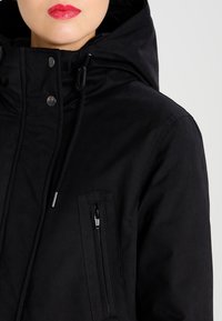 Zwarte hooded jack met een ritszak, aanpasbare trekkoord en drukknoopsluiting; heeft een soepele textuur en een minimalistisch ontwerp.